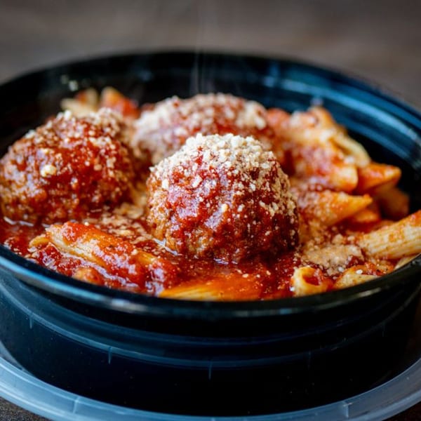 Steaming penne pasta with meatballs, marinara sauce, and grated cheese in a black takeout container.